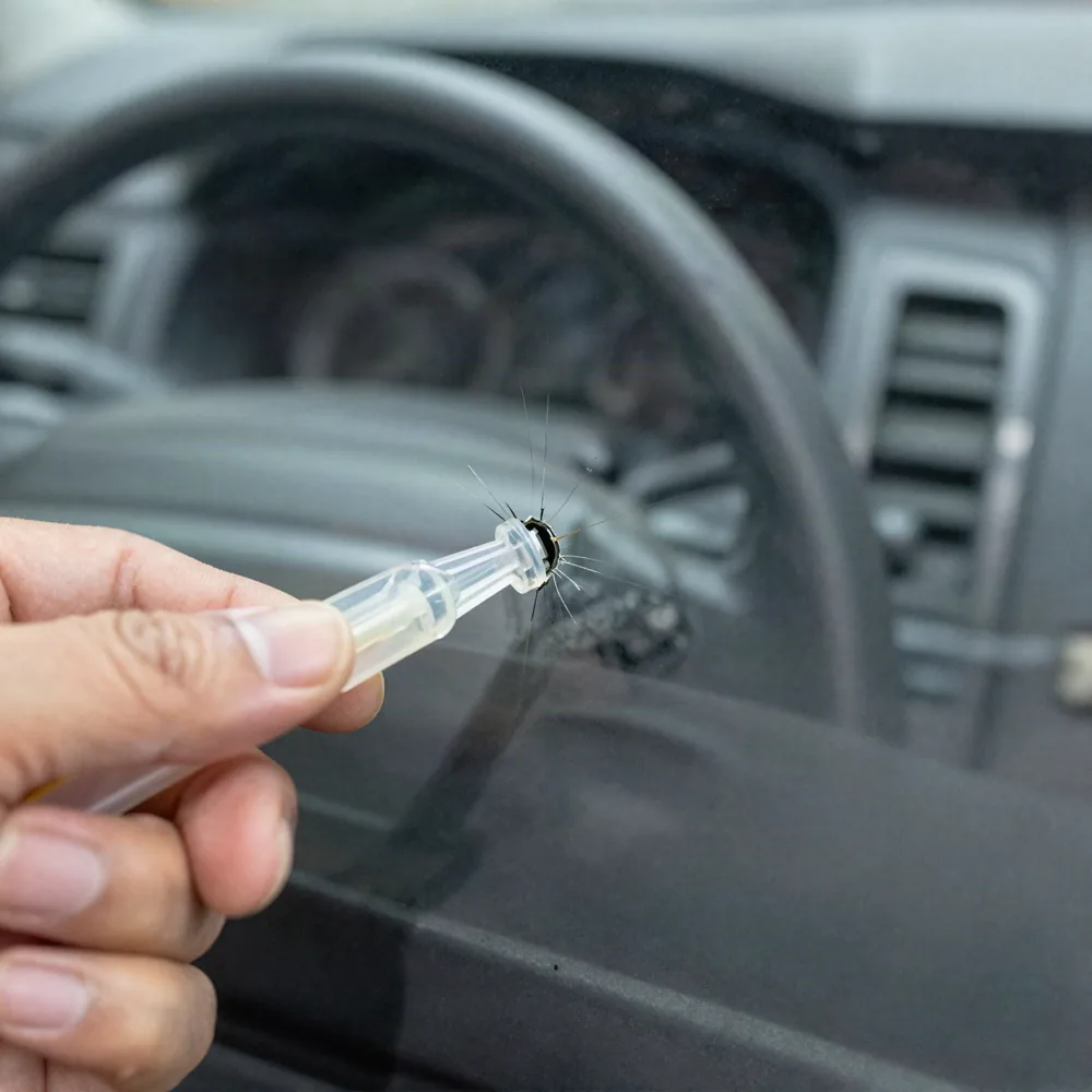Impact damage in a car windshield with crack legs radiating from the chip, a resin injector held over it and the dashboard visible behind the glass